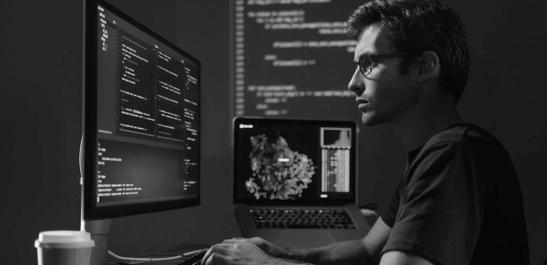 A developer at a dual-monitor desk reading code in a darkened room